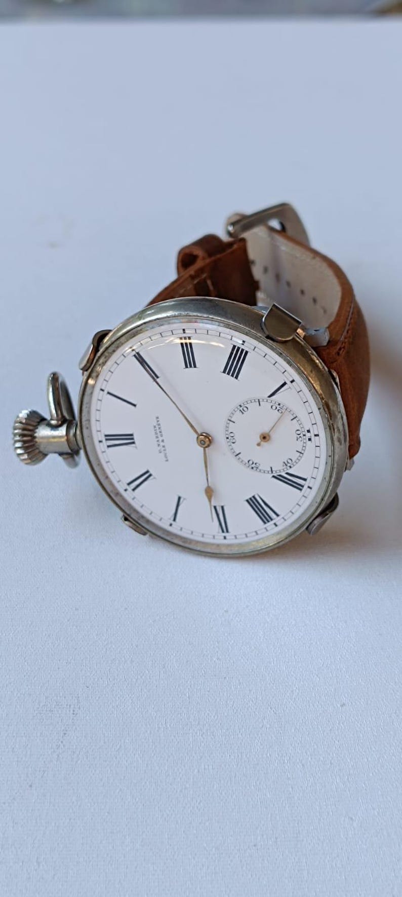 May include: A vintage pocket watch with a white face, black Roman numerals, and a small seconds dial. The watch has a silver-coloured case and a brown leather strap. The watch measures approximately 5 cm in diameter.