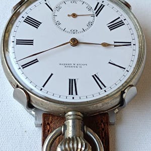 May include: A vintage U. Nardin Locle & Geneve pocket watch with a white face, black Roman numerals, and a subdial. The watch has a brown leather strap and a silver-tone case.