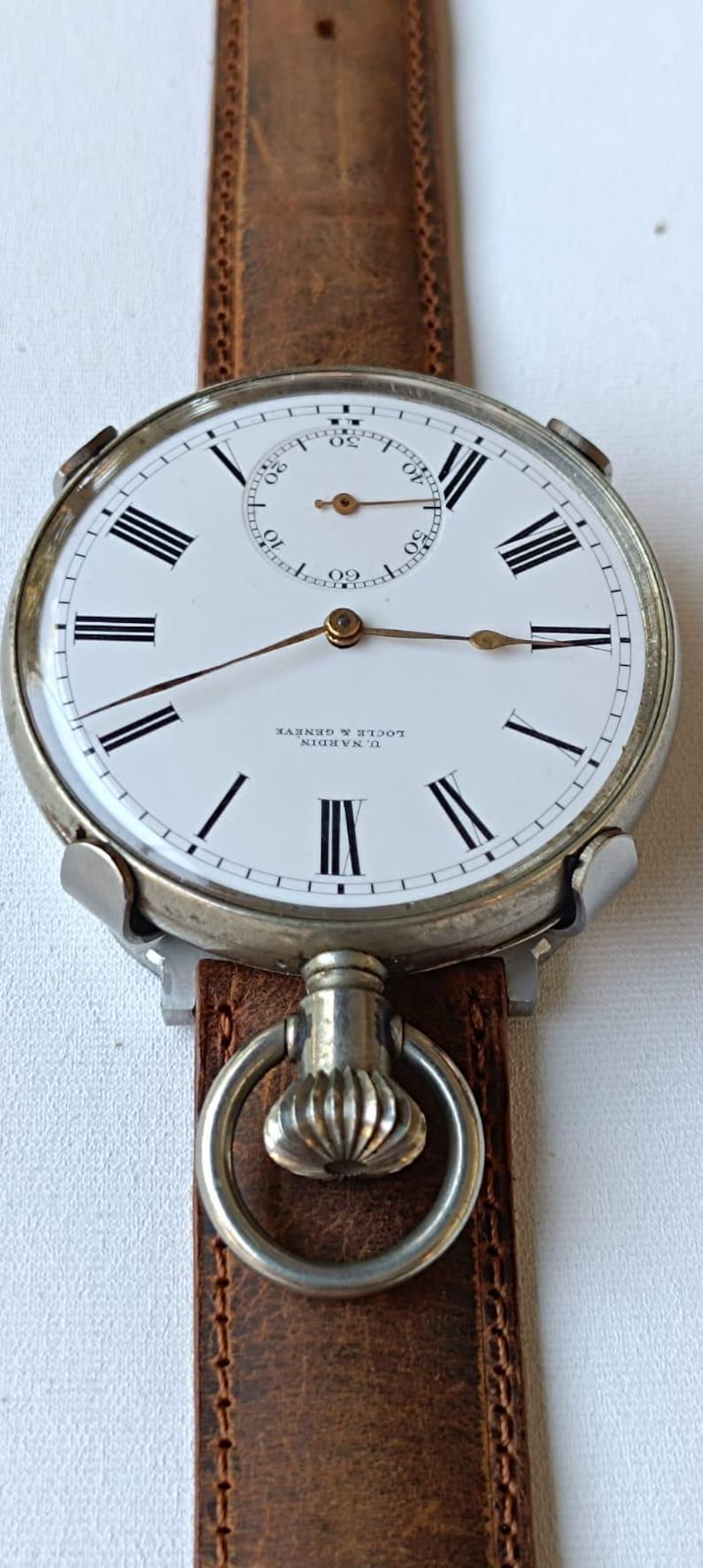 May include: A vintage U. Nardin Locle & Geneve pocket watch with a white face, black Roman numerals, and a subdial. The watch has a brown leather strap and a silver-tone case.