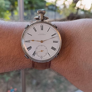 May include: A silver pocket watch with a white face, black Roman numerals, and a subdial. The watch has a brown leather strap. The watch face reads "H. WANDI, LOCLE & GENEVE".