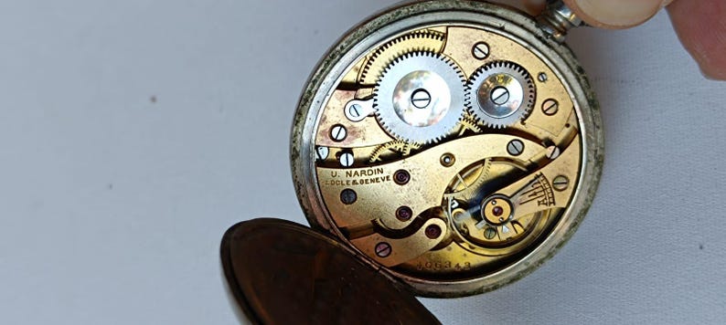 May include: An open antique pocket watch revealing its internal mechanisms. The gold-toned face displays silver gears and the inscription "U. NARDIN LOCLE & GENEVE". The silver-coloured watch case is partially visible.