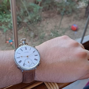 May include: A classic pocket watch with a white face, black Roman numerals, and a small seconds dial. The watch is on a brown leather strap. The watch is being worn on a wrist.