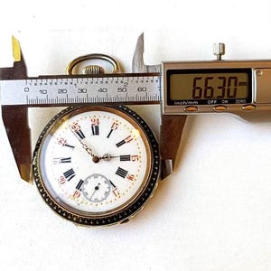 May include: A round pocket watch with a white face, black Roman numerals, and a sub-dial. The watch features a gold-tone case and a winding crown at the top. A digital caliper is measuring the watch, displaying a width of 66.30 millimeters.