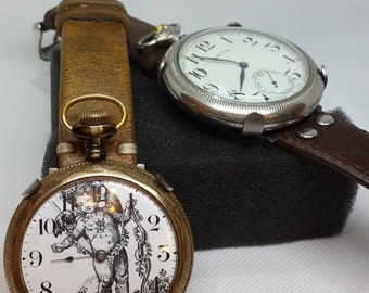 May include: Two antique pocket watches with leather straps. The watch on the left has a white face with a black and white image of a cherub. The watch on the right has a white face with black Roman numeral hour markers.