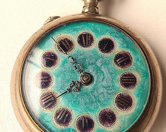 May include: Vintage pocket watch with a silver case and a turquoise face. The watch face has purple Roman numerals and gold detailing. The hands are black and decorative. The winding crown is gold-coloured.