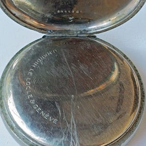 May include: An open, antique pocket watch with a silver-coloured case. The watch face is visible, with the words "U. Nardin Le Locle & Geneve" inscribed. The watch has a ring for attaching a chain.