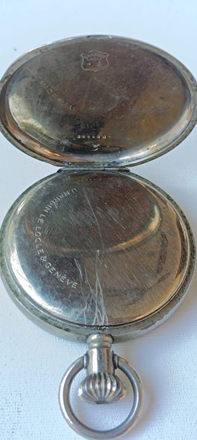 May include: An open, antique pocket watch with a silver-coloured case. The watch face is visible, with the words "U. Nardin Le Locle & Geneve" inscribed. The watch has a ring for attaching a chain.