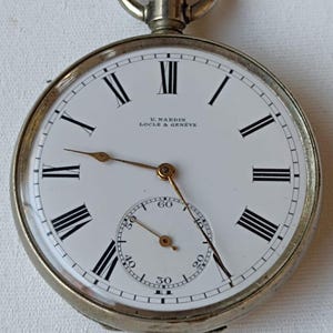 May include: A silver pocket watch with a white face and black Roman numerals. The watch has a subdial and the text "U. NARDIN LOCLE & GENEVE" is printed on the face. The watch has a crown and a loop.