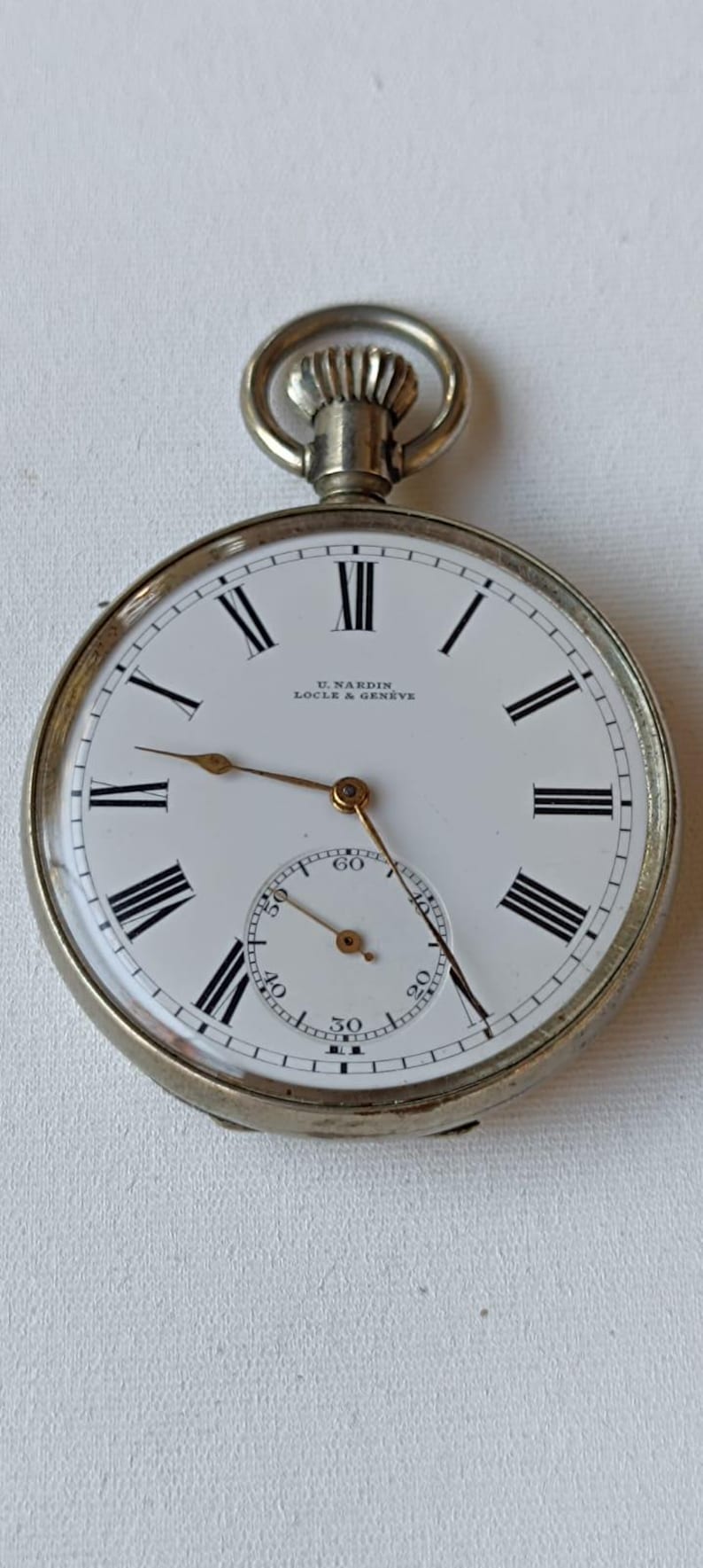 May include: A silver pocket watch with a white face and black Roman numerals. The watch has a subdial and the text "U. NARDIN LOCLE & GENEVE" is printed on the face. The watch has a crown and a loop.