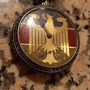 Op de afbeelding: Antieke zakhorloge met een zilverkleurige kast en een gedetailleerde wijzerplaat. De wijzerplaat toont een gouden adelaarslogo op een rode en groene achtergrond. Het horloge heeft een decoratieve kroon.
