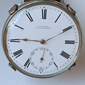 May include: A vintage U. Nardin Locle & Geneve pocket watch with a white face, black Roman numerals, and a subdial. The watch has a silver-tone case and a brown leather strap.