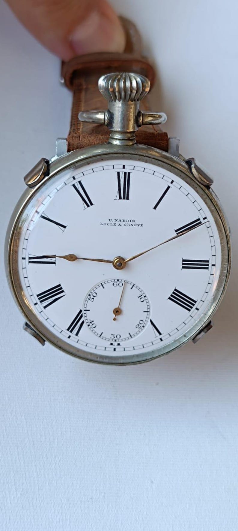 May include: A vintage U. Nardin Locle & Geneve pocket watch with a white face, black Roman numerals, and a subdial. The watch has a silver-tone case and a brown leather strap.