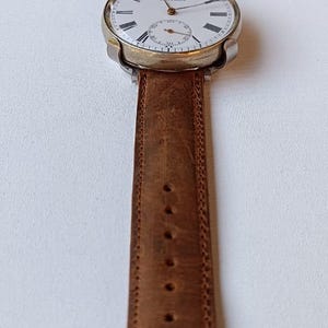 May include: A vintage wristwatch with a white face, black Roman numerals, and a subdial. The watch has a gold-coloured case and a brown leather strap. The strap is approximately 2 cm wide.