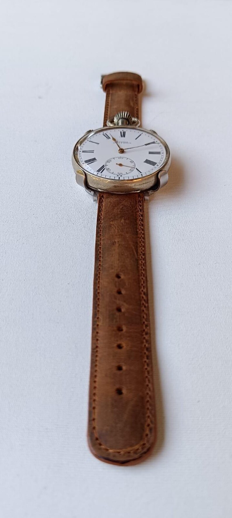 May include: A vintage wristwatch with a white face, black Roman numerals, and a subdial. The watch has a gold-coloured case and a brown leather strap. The strap is approximately 2 cm wide.