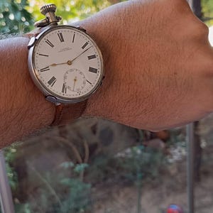 May include: A converted vintage pocket watch worn as a wristwatch. The watch features a white face with black Roman numerals and a subdial. It is attached to a brown leather strap.