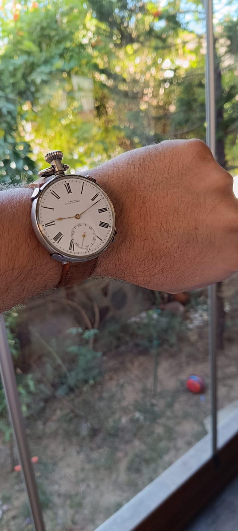 May include: A converted vintage pocket watch worn as a wristwatch. The watch features a white face with black Roman numerals and a subdial. It is attached to a brown leather strap.