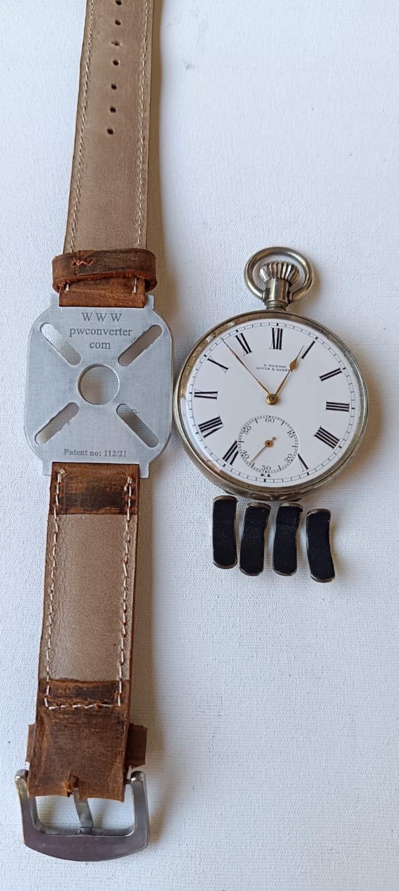 May include: A classic pocket watch with a white dial, black Roman numerals, and a seconds subdial. It is shown with a brown leather strap and a metal adapter with the text "www.pwconverter.com" and "Patent no: 112/21". Four black watch cushions are also present.
