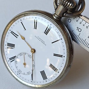 May include: A vintage pocket watch with a white face, black Roman numerals, and a subdial. The watch has a silver-coloured case and a crown at the top. The watch face reads "U. Nardin Locle & Geneve". A measuring tool is next to the watch.