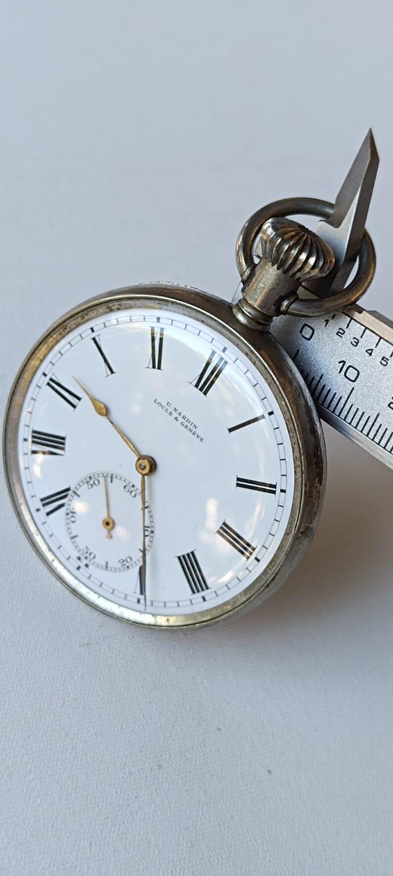 May include: A vintage pocket watch with a white face, black Roman numerals, and a subdial. The watch has a silver-coloured case and a crown at the top. The watch face reads "U. Nardin Locle & Geneve". A measuring tool is next to the watch.