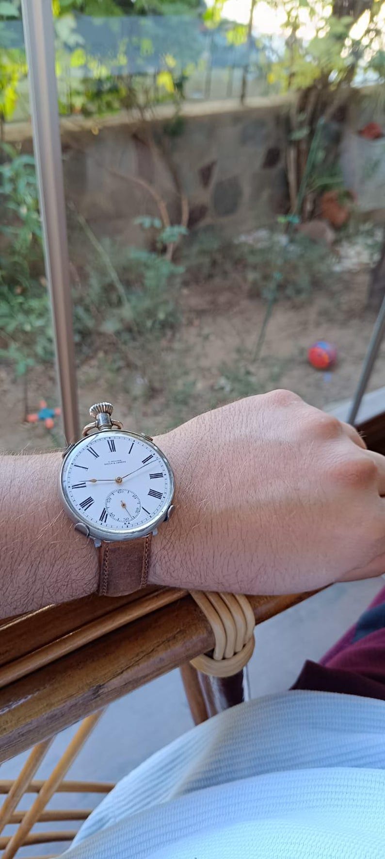 May include: A vintage pocket watch with a white face, black Roman numerals, and a smaller seconds dial. The watch is on a brown leather strap. The watch is being worn on a wrist.