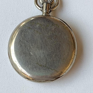 May include: A silver pocket watch with a circular face and a ring at the top. The watch has a brushed metal finish and a hinged back. The watch is on a white surface.