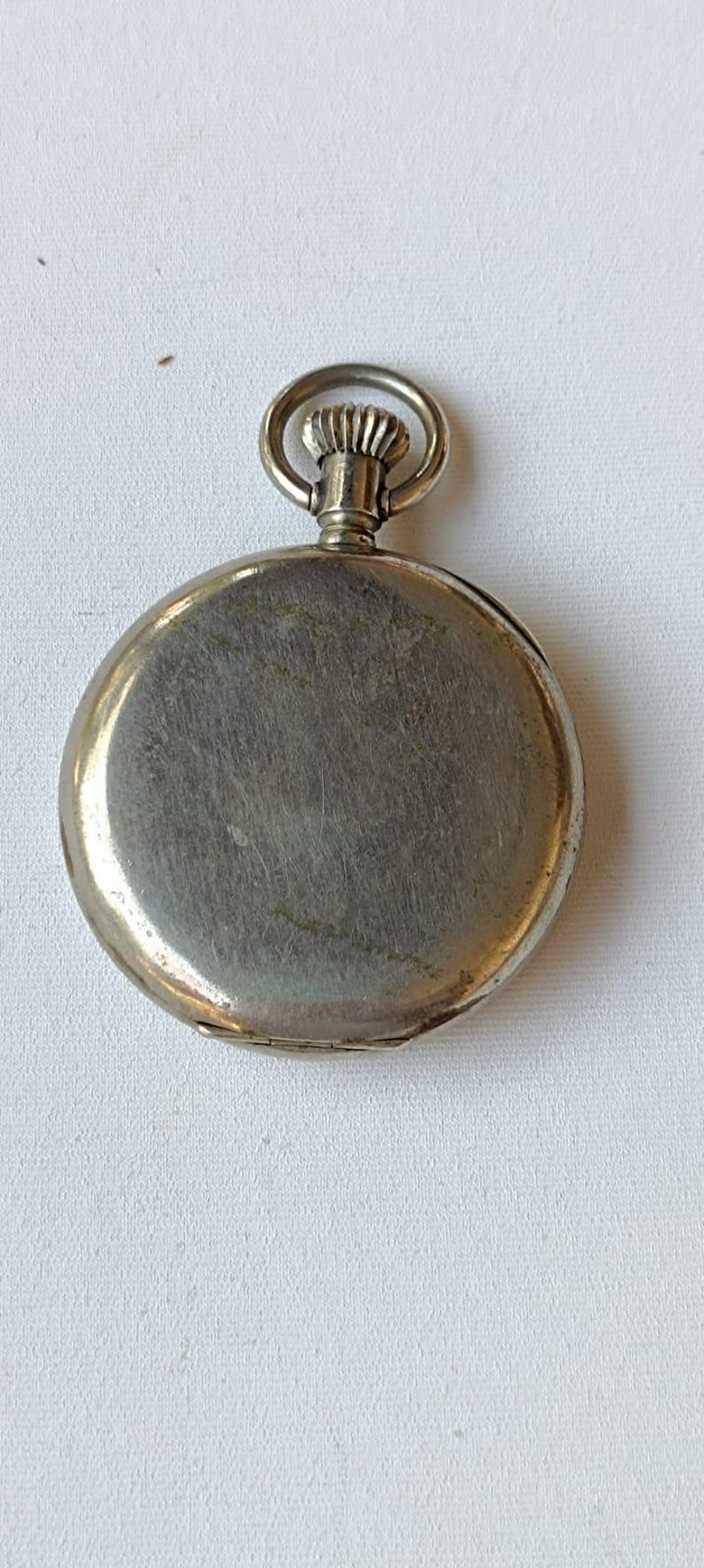 May include: A silver pocket watch with a circular face and a ring at the top. The watch has a brushed metal finish and a hinged back. The watch is on a white surface.