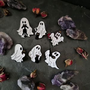 Könnte beinhalten: Satz mit sechs Emaille-Pins mit weißen Geistern, die schwarze Katzen halten. Die Geister haben schwarze Augen und sind schwarz umrandet. Die Katzen sind schwarz mit gelben Augen. Die Pins sind auf einer dunkelgrauen Oberfläche mit violetten Kristallen und getrockneten Rosenknospen angeordnet.