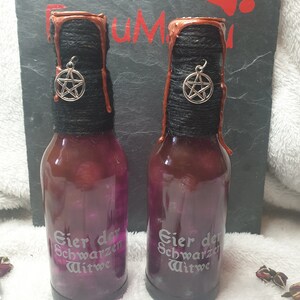 Könnte beinhalten: Zwei lilafarbene Glasflaschen mit schwarzem Garn und einem silbernen Pentagramm-Anhänger. Die Flaschen sind mit "Bier der Schwarzen Witwe" beschriftet, was "Bier der Schwarzen Witwe" bedeutet.