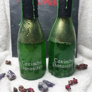 Könnte beinhalten: Zwei grüne Glasflaschen mit schwarzen Wachstropfen und goldenen Akzenten. Die Flaschen sind mit einer grünen Flüssigkeit gefüllt und tragen den Text "Toxische Parasiten" auf der Vorderseite.