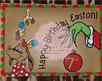 Pode incluir: Uma faixa de aniversário pintada à mão com uma rena de desenho animado e uma mão verde segurando um enfeite vermelho com o número 7. A faixa tem as palavras "Happy Birthday Easton!" em preto.