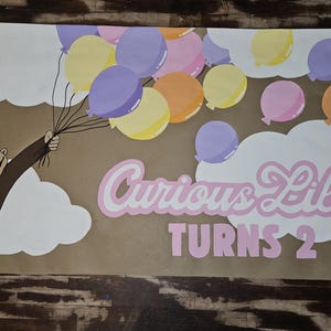 May include: A brown banner featuring a cartoon monkey holding balloons. The banner reads "Curious Lilah TURNS 2" in pink and white lettering. Balloons are yellow, purple, pink, and orange.