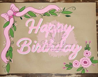 May include: A hand-painted banner with the words "Happy Birthday" in pink and white, adorned with pink ribbons, green leaves, and pink flowers. The banner is on a brown background.