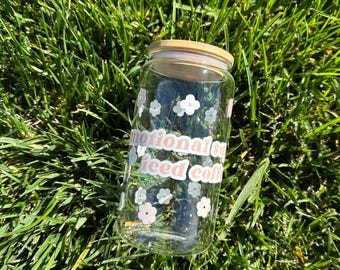 May include: A clear glass jar with a wooden lid and a pink and white sticker that says "emotional support iced coffee" with white flowers on the jar.