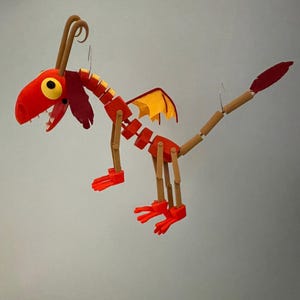 May include: A red and yellow articulated dragon toy with a segmented body, wings, and tail. The dragon has a red head with large yellow eyes and a toothy grin. The wings are yellow and orange. The toy is suspended by a thin wire.