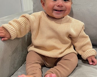 Puede incluir: Un bebé con un suéter de punto beige y leggings de canalé marrones está sentado en un sofá gris claro. El bebé sonríe y mira a la cámara.