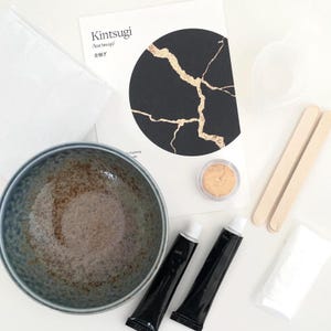 May include: Kintsugi repair kit featuring a ceramic bowl, black tubes, gold powder, and wooden stirrers. The kit includes a card with the word "Kintsugi" and a black circle design with gold cracks, representing the art of repairing broken pottery.