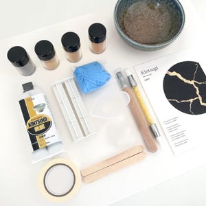 May include: Kintsugi repair kit with various components. Includes glue, powders, brushes, a bowl, tape, and a guide. The kit features a blue bowl and a sheet with a black circle and gold cracks.