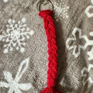 K&ouml;nnte beinhalten: Ein roter geflochtener Schl&uuml;sselanh&auml;nger mit einem silberfarbenen Karabiner und Schl&uuml;sselring. Der Schl&uuml;sselanh&auml;nger besteht aus einem dicken, strukturierten Garn und hat eine Quaste am Ende. Der Hintergrund ist ein weicher, grauer Stoff mit Schneeflockenmuster.