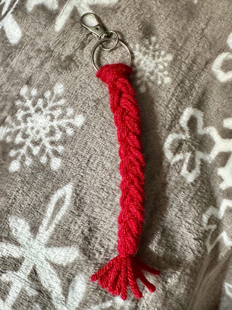 K&ouml;nnte beinhalten: Ein roter geflochtener Schl&uuml;sselanh&auml;nger mit einem silberfarbenen Karabiner und Schl&uuml;sselring. Der Schl&uuml;sselanh&auml;nger besteht aus einem dicken, strukturierten Garn und hat eine Quaste am Ende. Der Hintergrund ist ein weicher, grauer Stoff mit Schneeflockenmuster.