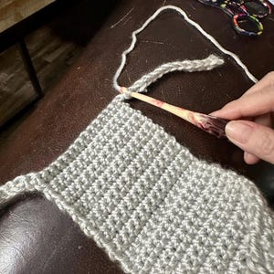 May include: Close-up of a crochet project in progress. A light gray crocheted piece is being worked on with a wooden crochet hook. Floral scissors and a ball of yarn are also visible.