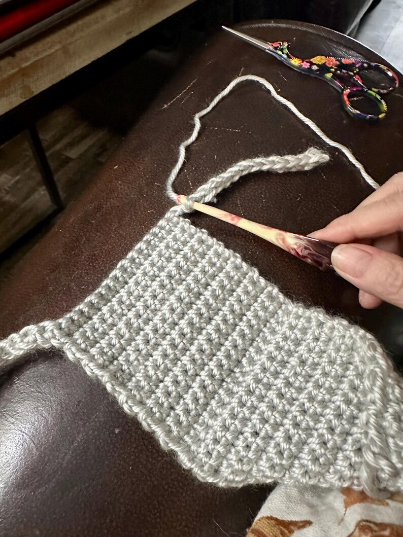 May include: Close-up of a crochet project in progress. A light gray crocheted piece is being worked on with a wooden crochet hook. Floral scissors and a ball of yarn are also visible.