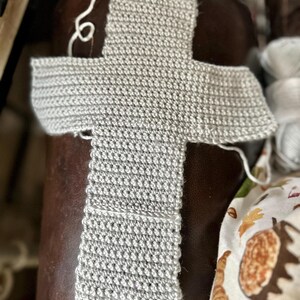 May include: A light gray, unfinished crochet project in a cross shape. The handmade piece is made of a textured yarn and sits on a brown leather surface. White yarn is visible, suggesting ongoing work. The project is likely a scarf or shawl.