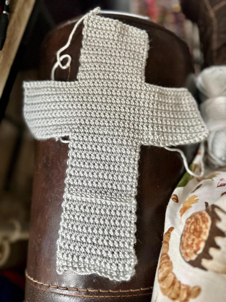 May include: A light gray, unfinished crochet project in a cross shape. The handmade piece is made of a textured yarn and sits on a brown leather surface. White yarn is visible, suggesting ongoing work. The project is likely a scarf or shawl.