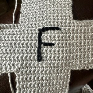 May include: Close-up of a crocheted gray fabric with a black embroidered letter "F". The fabric is made of a textured yarn and appears to be part of a larger project. The stitches are clearly visible.