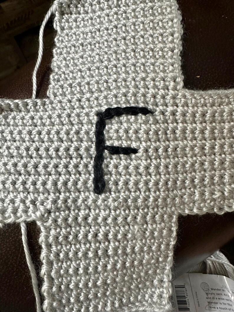 May include: Close-up of a crocheted gray fabric with a black embroidered letter "F". The fabric is made of a textured yarn and appears to be part of a larger project. The stitches are clearly visible.