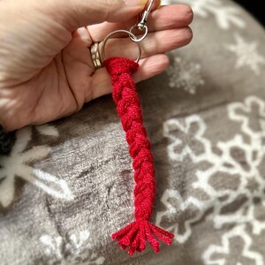 K&ouml;nnte beinhalten: Ein roter geflochtener Schl&uuml;sselanh&auml;nger mit einem silberfarbenen Karabiner und Schl&uuml;sselring. Der Schl&uuml;sselanh&auml;nger besteht aus einem dicken, gewebten Material und hat eine Quaste am Ende. Der Hintergrund ist eine weiche, graue Decke mit einem Schneeflockenmuster.