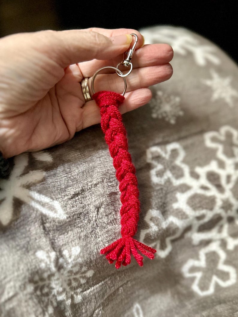 K&ouml;nnte beinhalten: Ein roter geflochtener Schl&uuml;sselanh&auml;nger mit einem silberfarbenen Karabiner und Schl&uuml;sselring. Der Schl&uuml;sselanh&auml;nger besteht aus einem dicken, gewebten Material und hat eine Quaste am Ende. Der Hintergrund ist eine weiche, graue Decke mit einem Schneeflockenmuster.