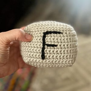May include: A crocheted, beige cube with a large black letter "F" on one side. The handmade item is held in a person's hand, showcasing the texture of the yarn and the simplicity of the design. The background is blurred.
