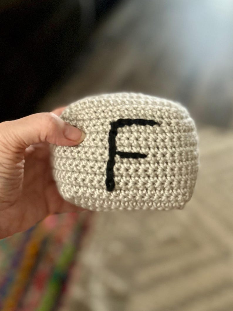 May include: A crocheted, beige cube with a large black letter "F" on one side. The handmade item is held in a person's hand, showcasing the texture of the yarn and the simplicity of the design. The background is blurred.