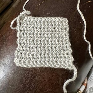 May include: A small, square swatch of light gray crochet fabric. The stitches are tightly woven, creating a textured surface. White yarn extends from the top and right side of the swatch, indicating the beginning and end of the work. The swatch is on a brown surface.